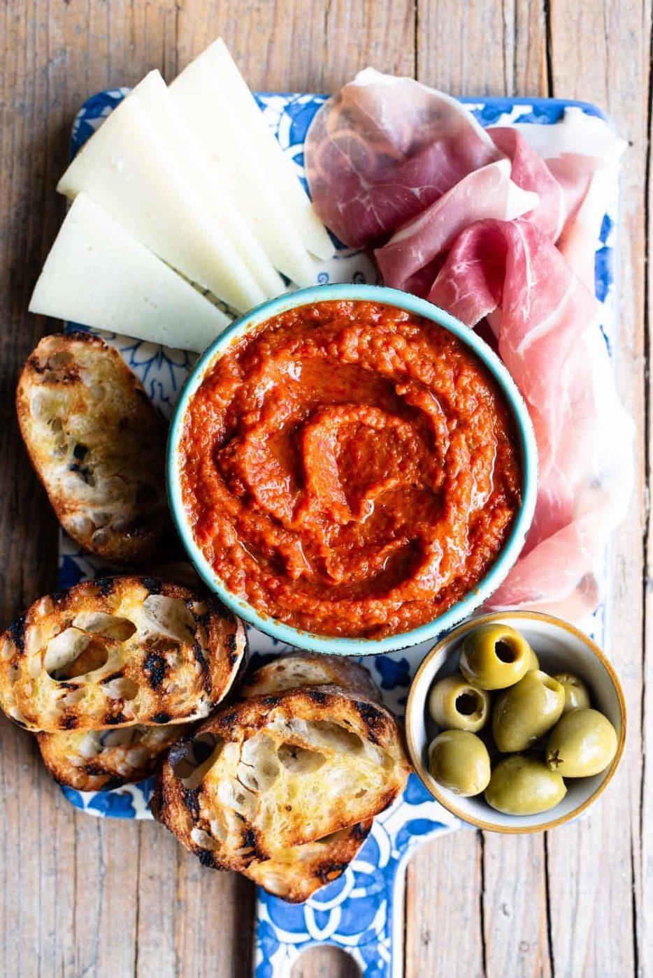 An overhead shot of Ajvar spread in a blue bowl sitting on a serving board with cured meats, cheese and olives.