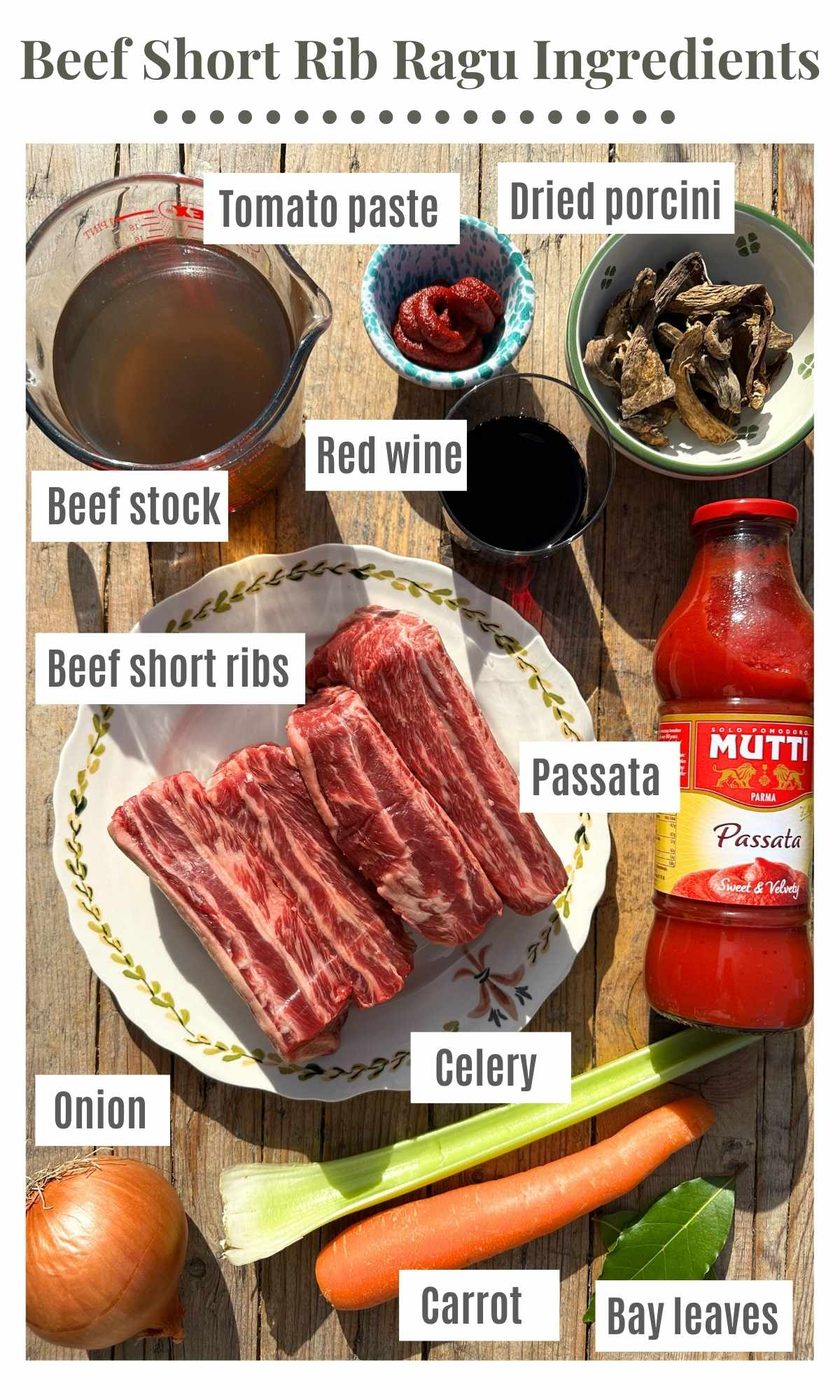 Beef short rib ragu ingredients arranged on a rustic wooden surface, including raw beef short ribs, onion, celery, carrot, beef stock, tomato paste, red wine, dried porcini, passata, and bay leaves, all labeled