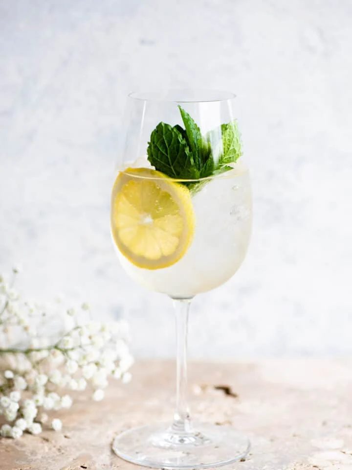 A Hugo spritz cocktail on a light coloured worktop with lemon and mint.