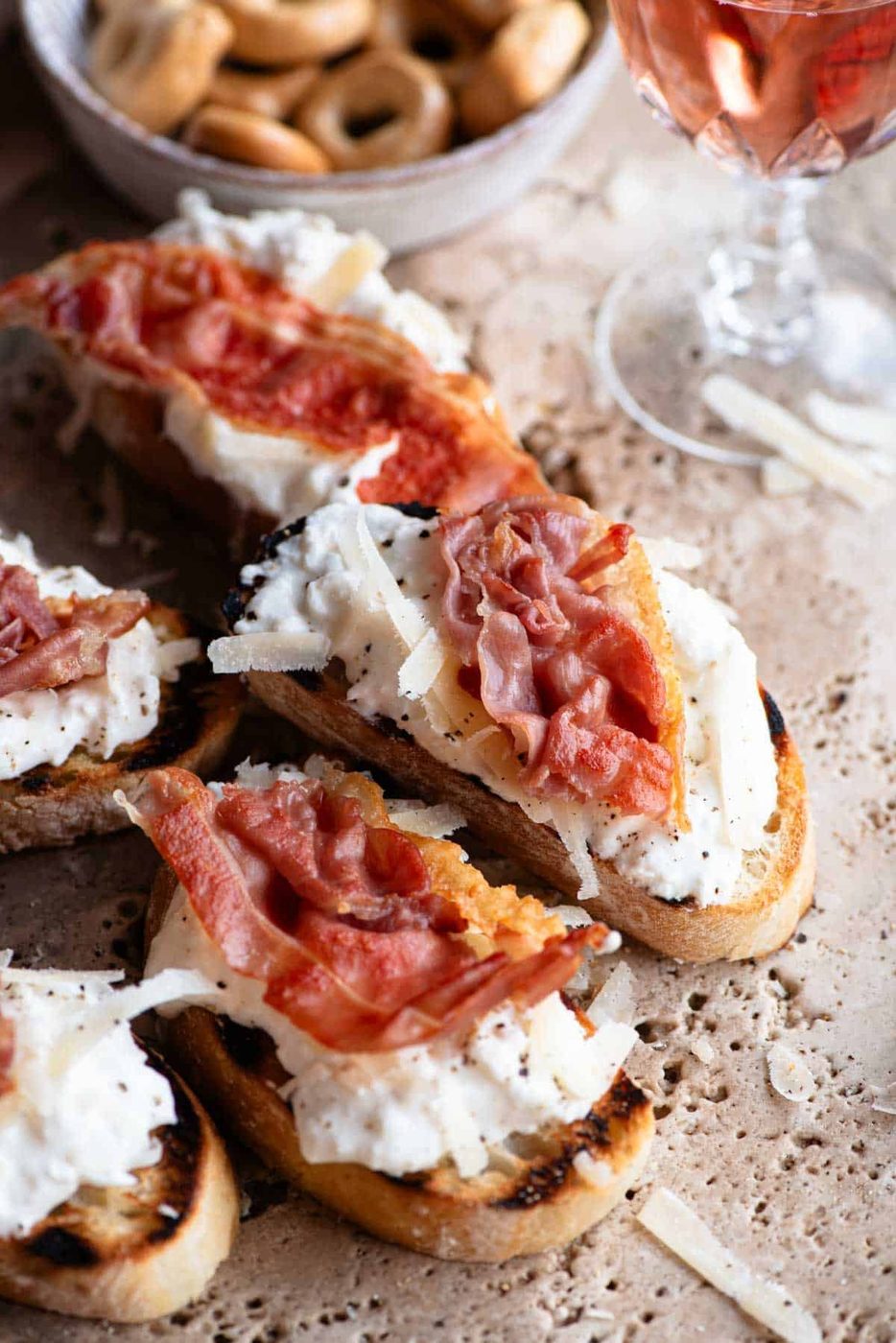 A close up of a slice of crostini topped with burrata, parmigiano reggiano and crispy prosciutto.