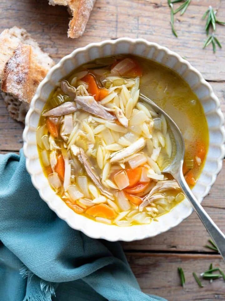 An overhead shot of a bowl of lemon chicken orzo soup on a wooden surface with crusty bread at the side.