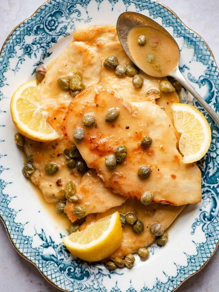 An overhead shot of chicken piccata on a blue serving plate with lemon wedges and capers