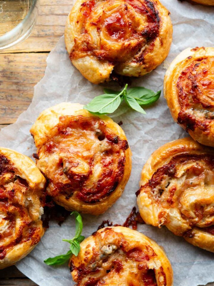 An overhead shot of pizza pinwheels on a wooden board with a basil leaf and glass of wine at the side.