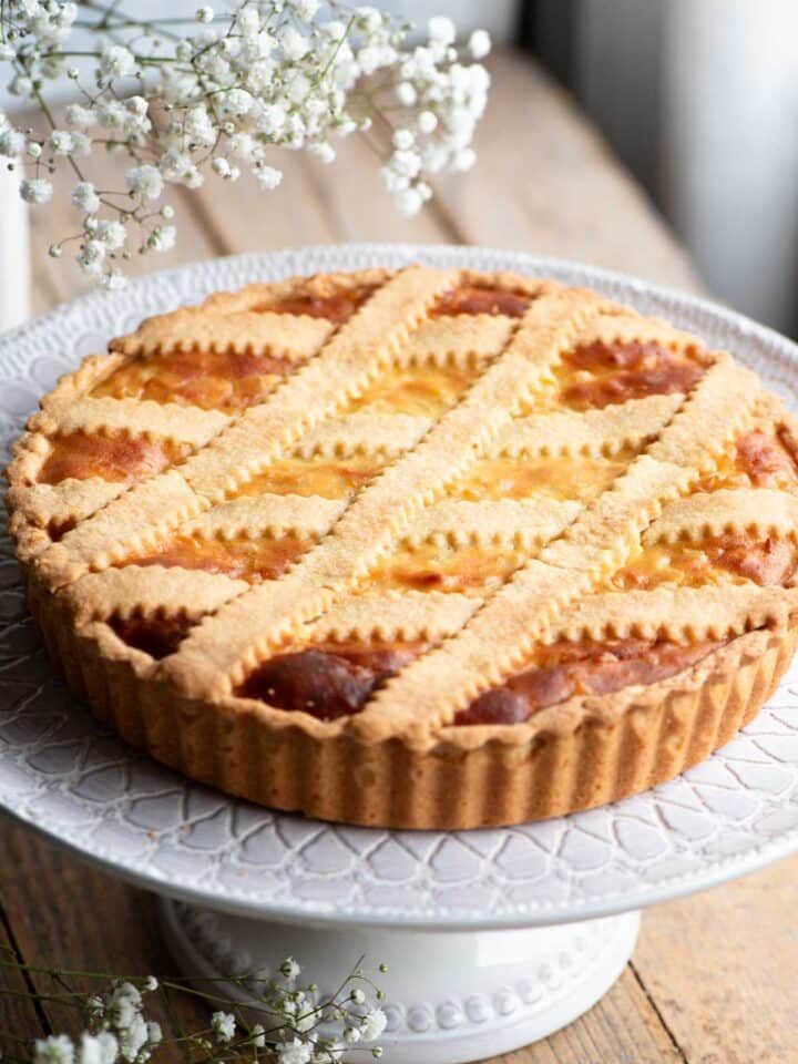 Pastiera Napoletana on a cake stand sitting on a wooden surface with white flowers at the side.