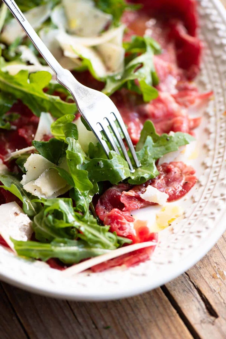 A close up of beef carpaccio, arugula and Grana Padano cheese on a plate with a fork.