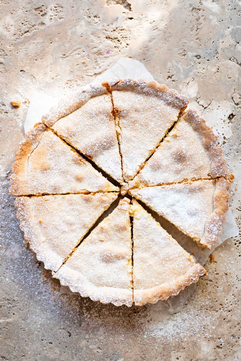 A crostata cut into 8 slices and dusted in powdered sugar sitting on a travertine surface.
