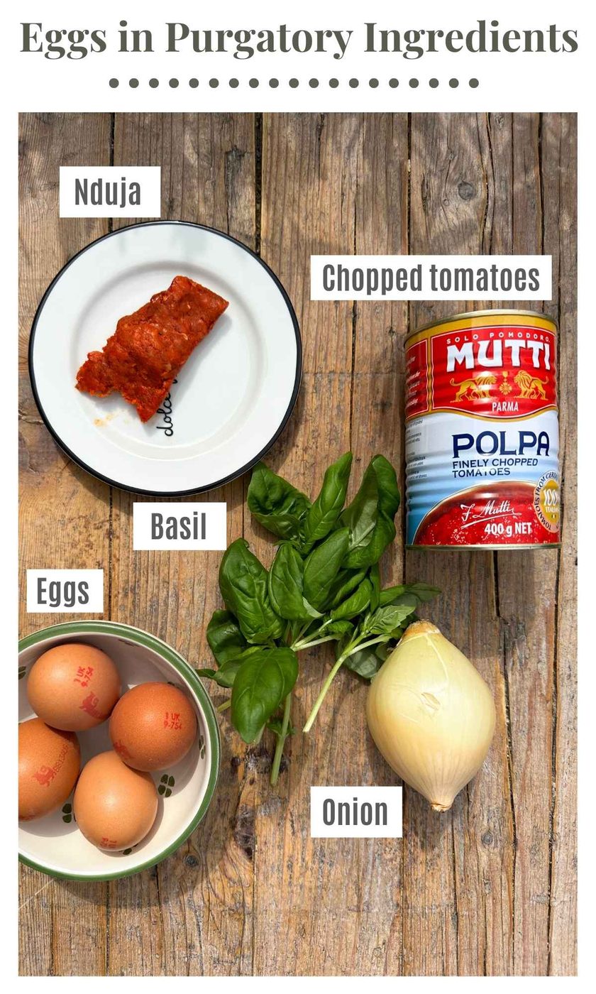 The ingredients to make spicy nduja eggs in purgatory on a rustic wooden surface. The ingredients are labelled: Nduja, chopped tomatoes, onion, basil, eggs.