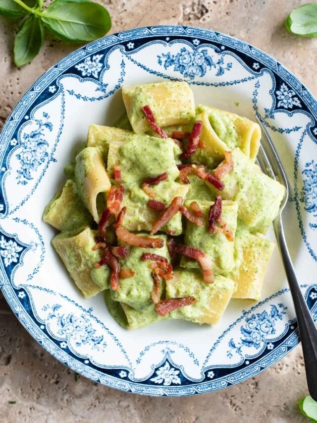 A creamy zucchini pasta in a blue and white bowl topped with crispy pancetta sitting on a travertine surface.