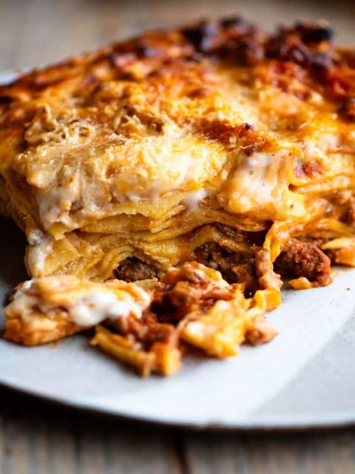 One slice of lamb lasagna on a plate sitting on a wooden surface.