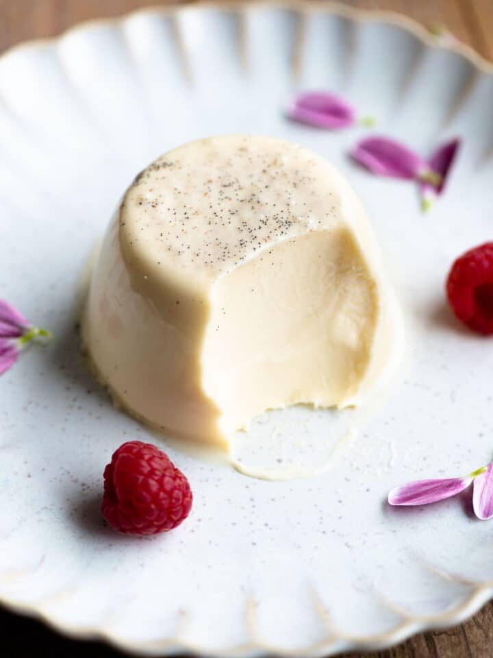 A vanilla panna cotta on a plate with a bite out and raspberries at the side.