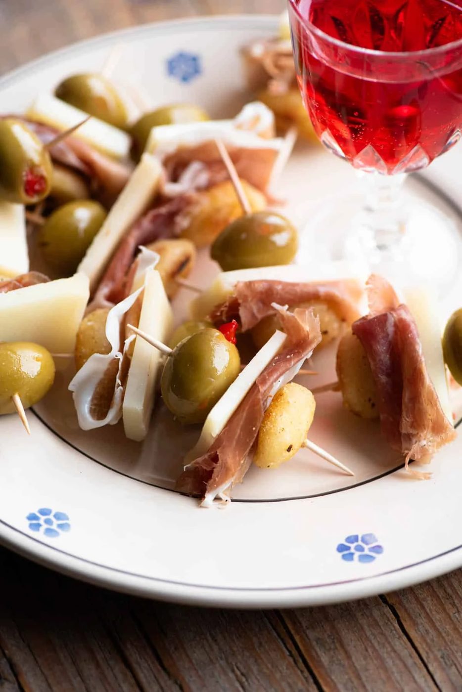 A close up of a fried gnocchi appetizer with pecorino cheese, prosciutto and an olive sitting on a plate with a red aperitivo drink.