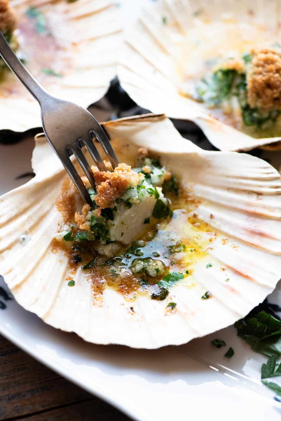 A close up of one scallop in a shell with parsley butter and a fork inserted into it.