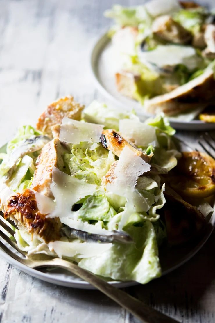 A side shot of a grilled chicken caesar salad on a white plate topped with shavings of parmesan cheese