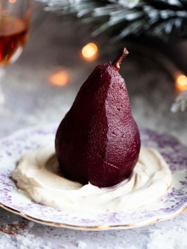 A red wine poached pear sitting on a bed of Amaretto whipped cream on a small plate, the background is Christmassy.