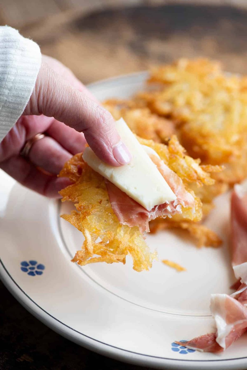 A close up of a hand holding a potato fritter topped with cured meat and cheese.