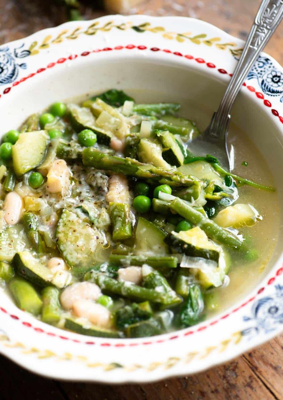 A close up of a green vegetable soup in a patterned bowl with asparagus, peas, zucchini and green beans.