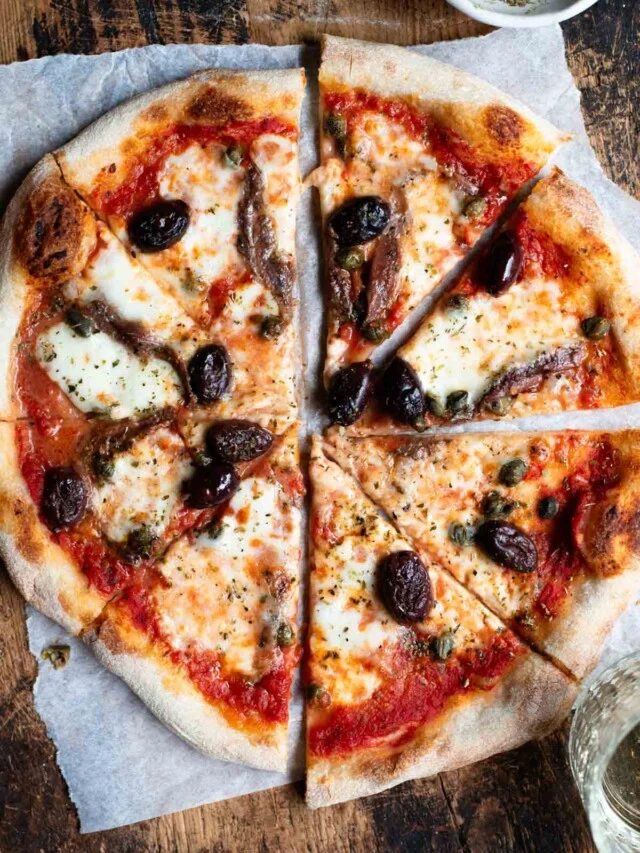 An overhead shot of a pizza cut into slices on a rustic wooden board. The pizza is topped with olives, anchovies and capers.