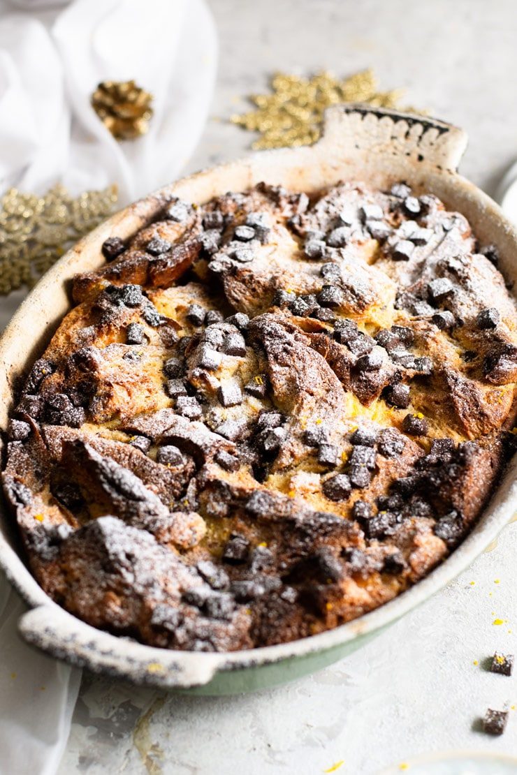 A close up of a panettone bread pudding in a rustic oval baking dish topped with chocolate chips and powdered sugar