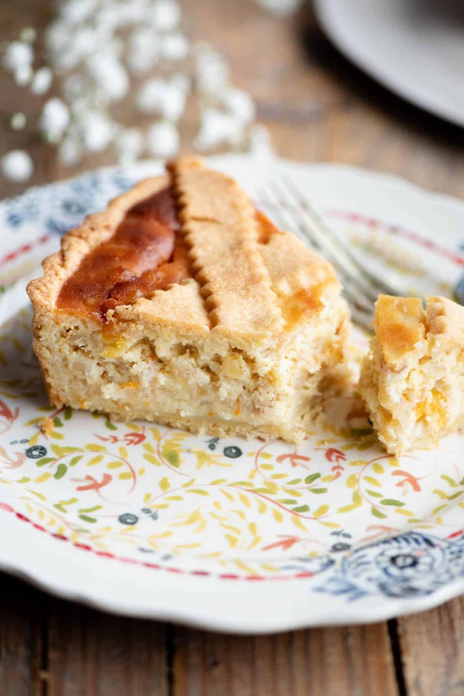 A slice of Pastiera Napoletana on a patterned plate with white flowers in the background.