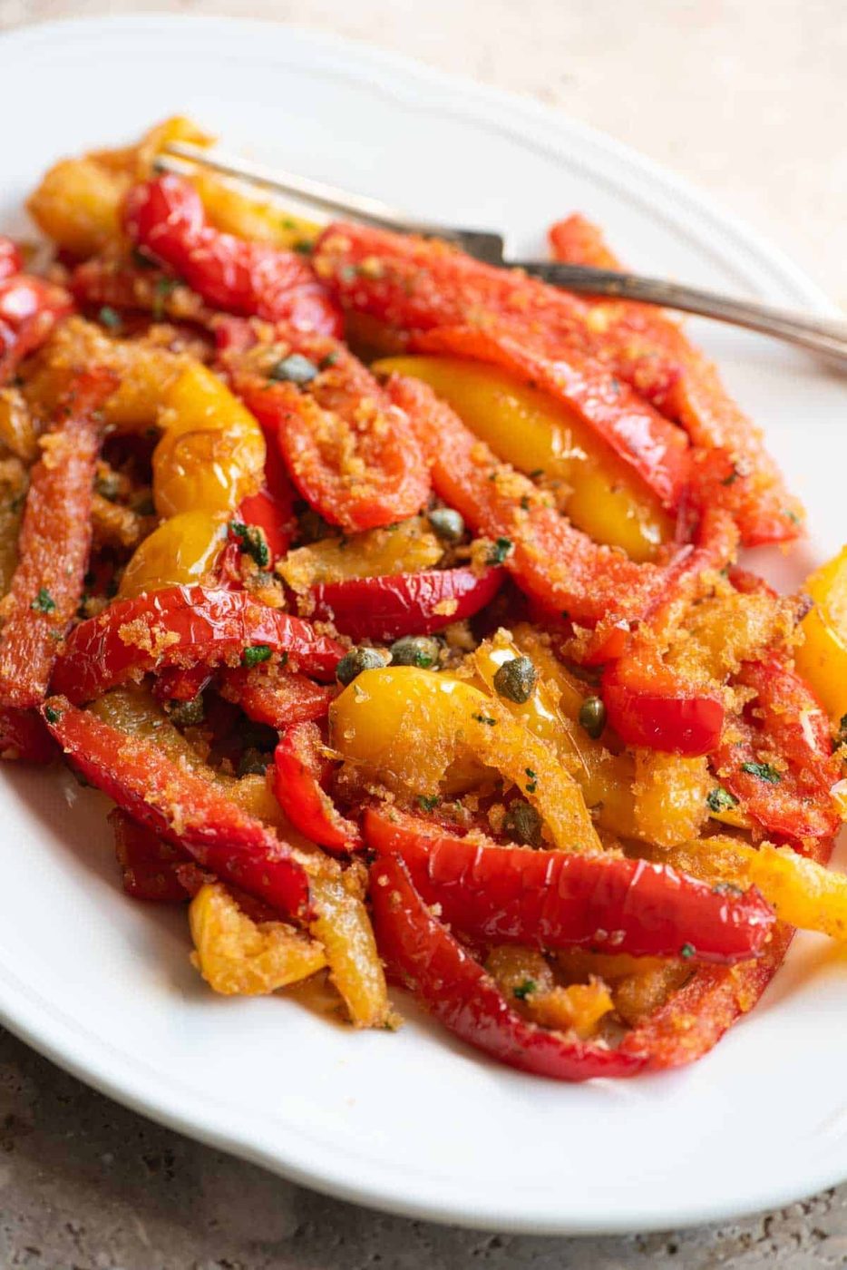 A close up side shot of mixed red and yellow peppers on a white servings dish with capers and breadcrumbs.