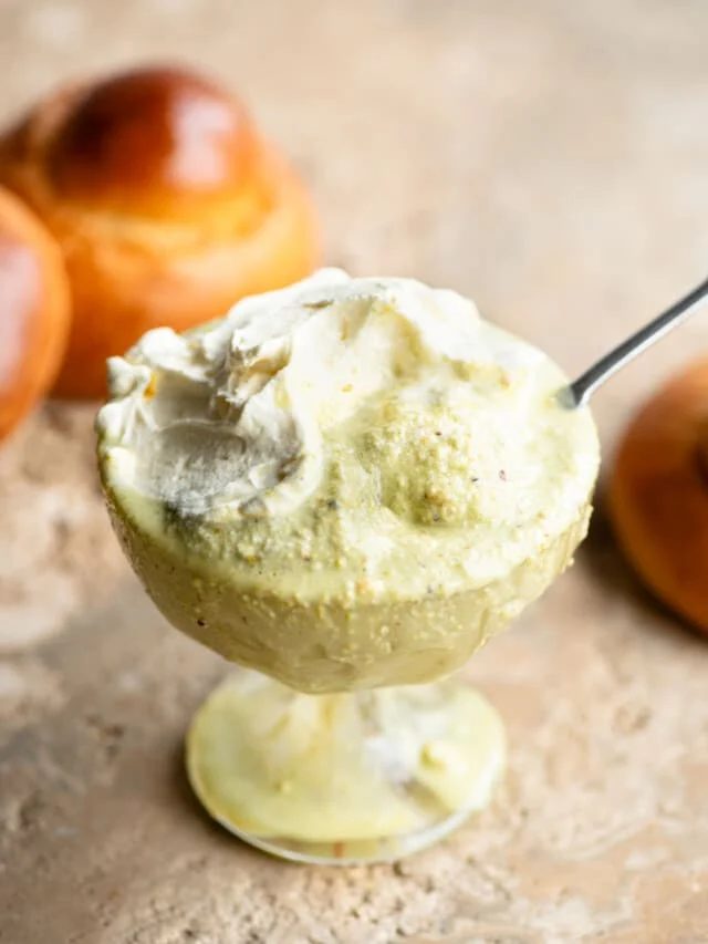 A close up of pistachio granita in a glass serving bowl topped with whipped cream and a brioche bun behind it.