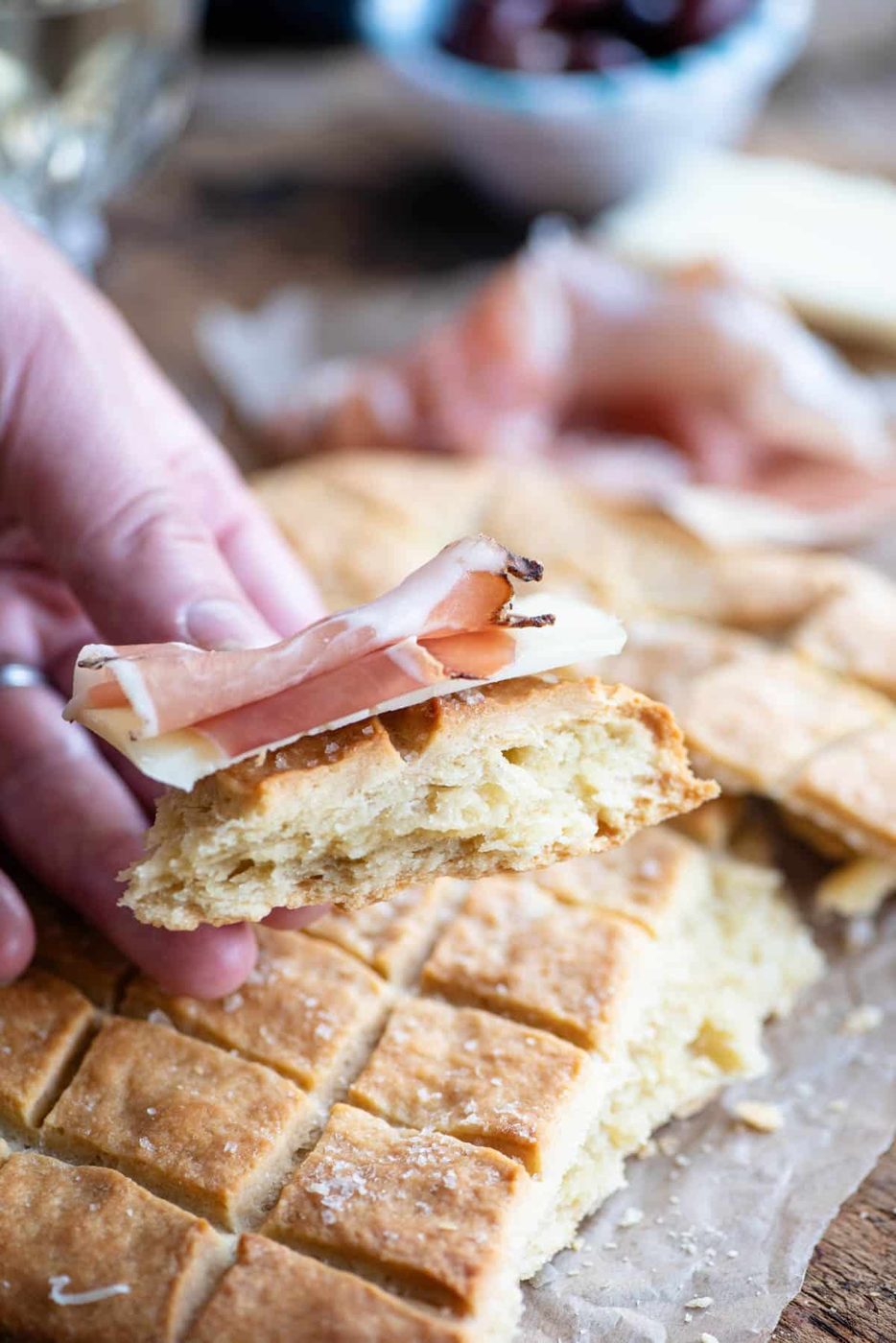 A close up of a hand holding a piece if Pizza Scima with cured meat.
