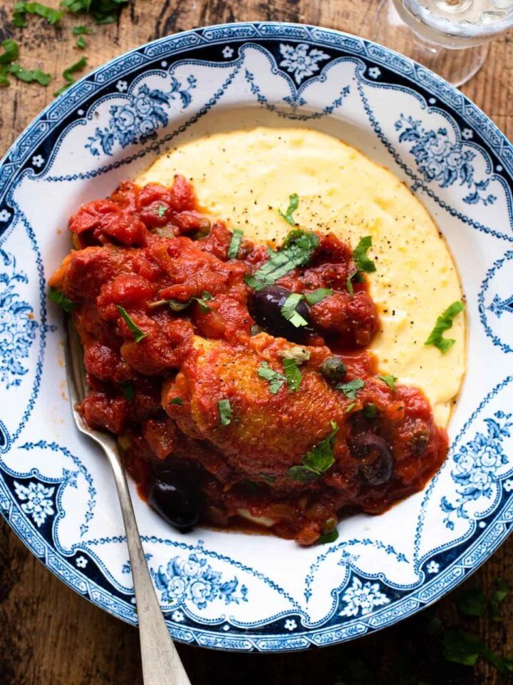 Chicken in Puttanesca sauce in a bowl with polenta sitting on a wooden surface.
