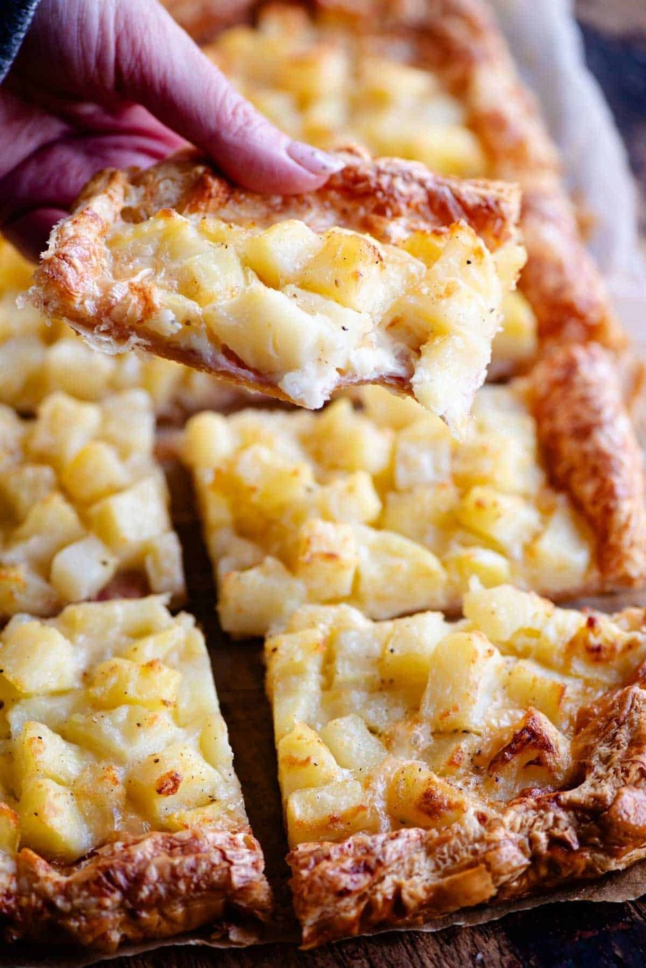 A close up of a hand holding a slice of puff pastry and potato tart.