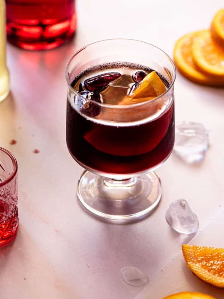 A red wine spritz (prampolini) on a light background with slices of orange, ice and campari at the side.