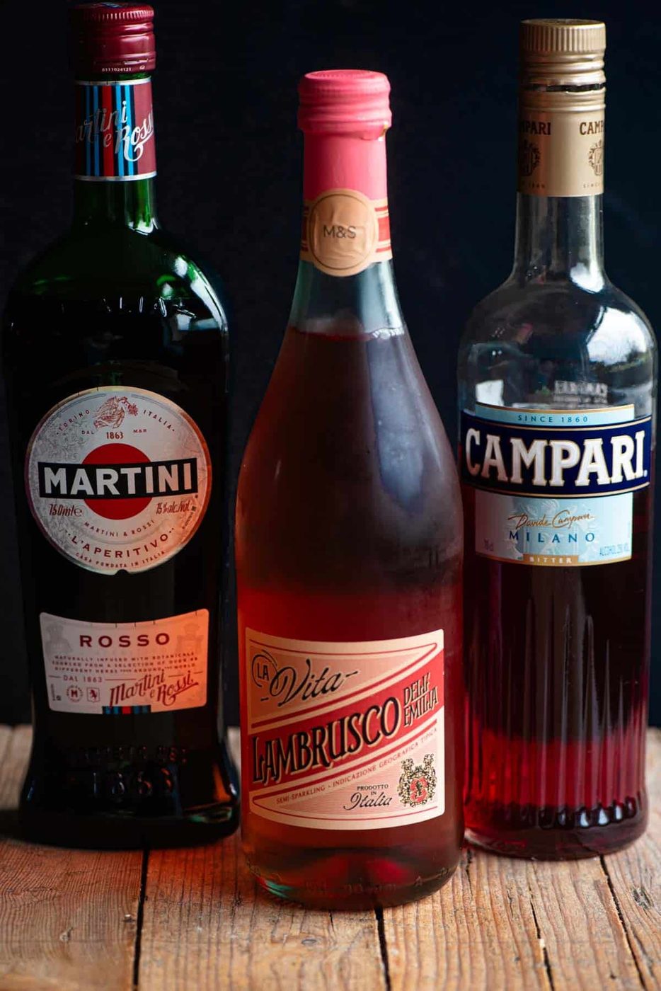 A bottle of Martini rosso, Lambrusco and Campari sitting on a wooden table with a black background.