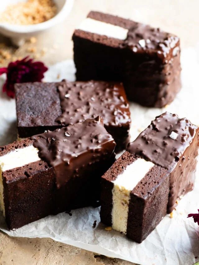 Four chocolate ice cream sandwiches with vanilla ice cream in the middle and dipped in chocolate and hazelnuts on a travertine surface.