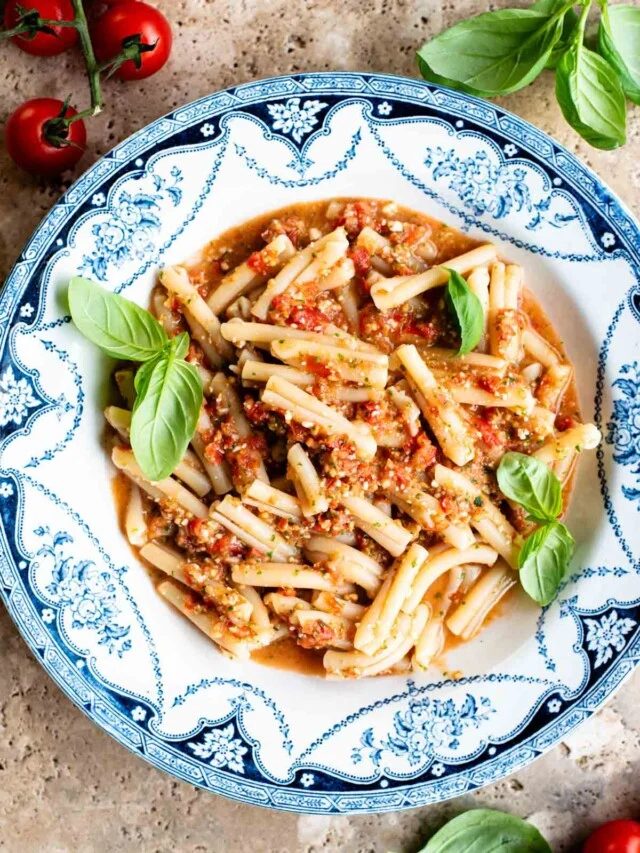 A blue and white vintage bowl with pasta in a fresh tomato sauce topped with basil. The bowl is on a travertine surface with cherry tomatoes and basil around.