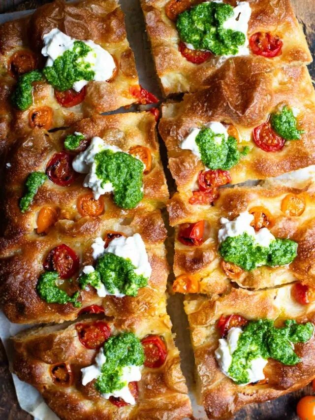 An overhead shot of a focaccia cut into 8 slices and topped with cherry tomatoes, pesto and burrata.