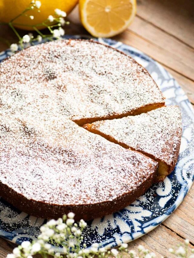 A white torta caprese on a blue ceramic board with fresh lemons behind it and small white flowers around.