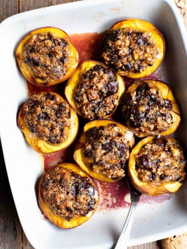 An overhead shot of baked peaches stuffed with amaretti cookies and chocolate in a white baking dish on a rustic wooden background.