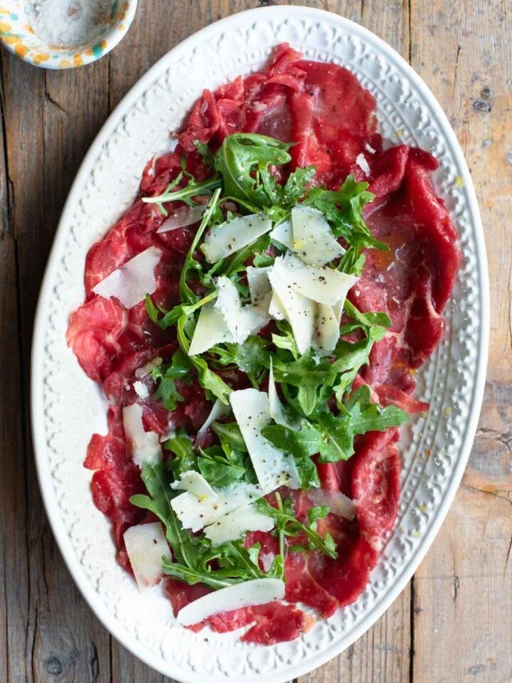 An overhead shot of beef carpaccio on a white oval serving plate topped with arugula and Grana Padano cheese.