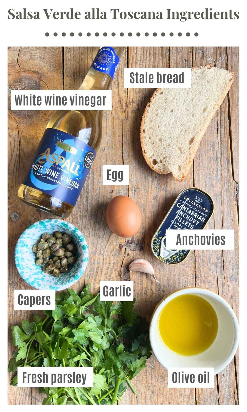 All the ingredients needed to make a Tuscan salsa verde on a wooden board. The text labels read; white wine vinegar, stale bread, garlic, anchovies, egg, capers, parsley, olive oil.