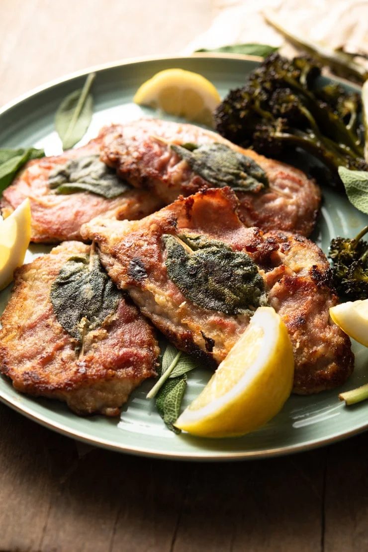 A side shot of Veal Saltimbocca, plus Prosciutto and Sage on a plate with lemon wedges