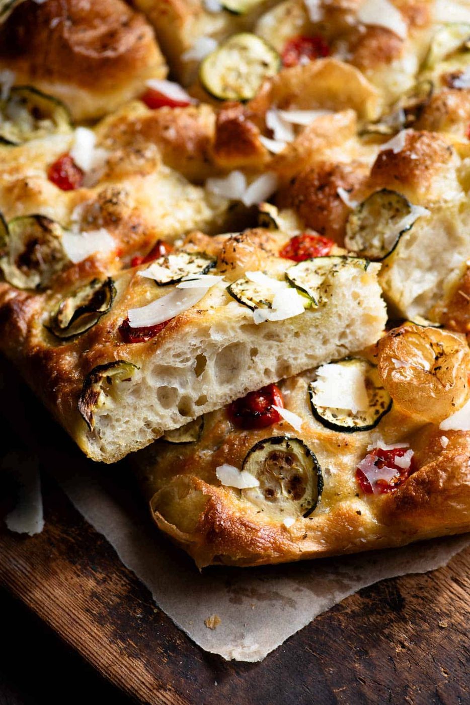 A side shot of a slice of focaccia showing the light and airy texture. It's topped with zucchini, tomatoes and pecorino on a rustic wooden board.