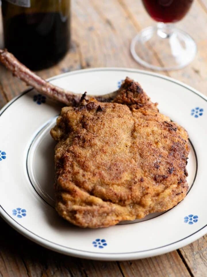 A breaded veal chop sitting on a plate with a glass and bottle of red wine in the background.