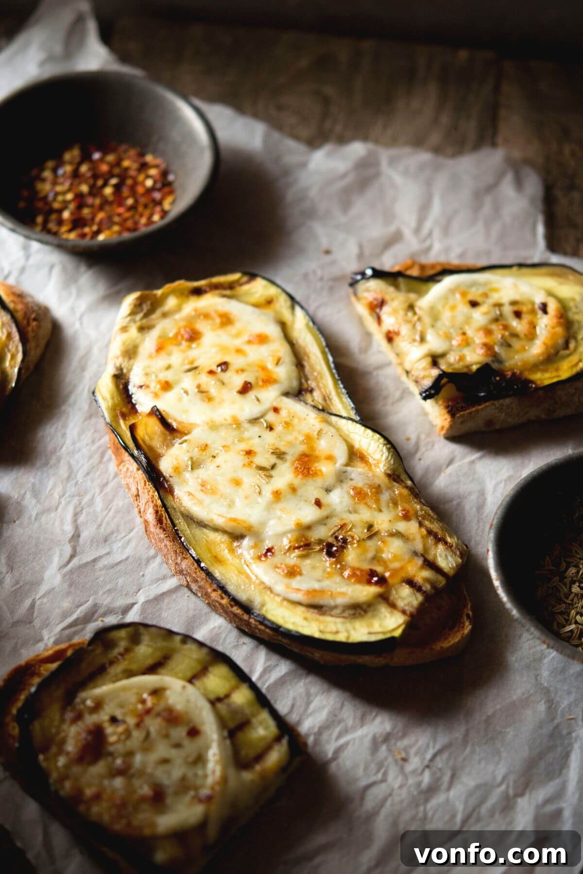 A slice of eggplant bruschetta in a rustic setting topped with melted mozzarella, red pepper flakes and fennel seeds.