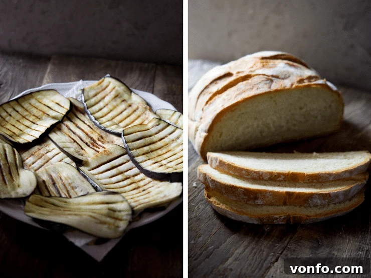 Grilled eggplant slices with distinctive char marks are arranged on a plate next to a loaf of crusty Italian bread, ready for assembly.