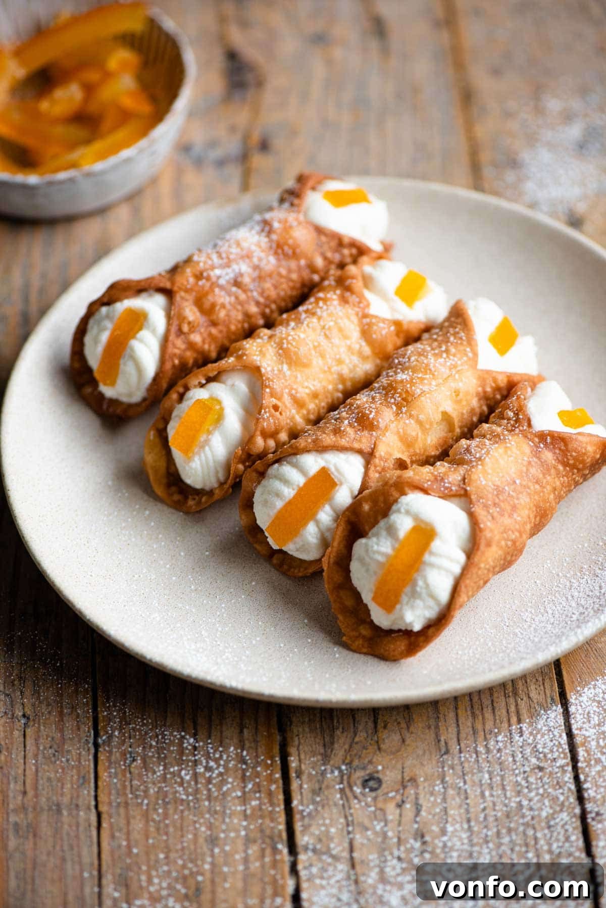 Four golden-brown cannoli on a white plate, generously filled with creamy ricotta and garnished with vibrant candied orange slices at each end.