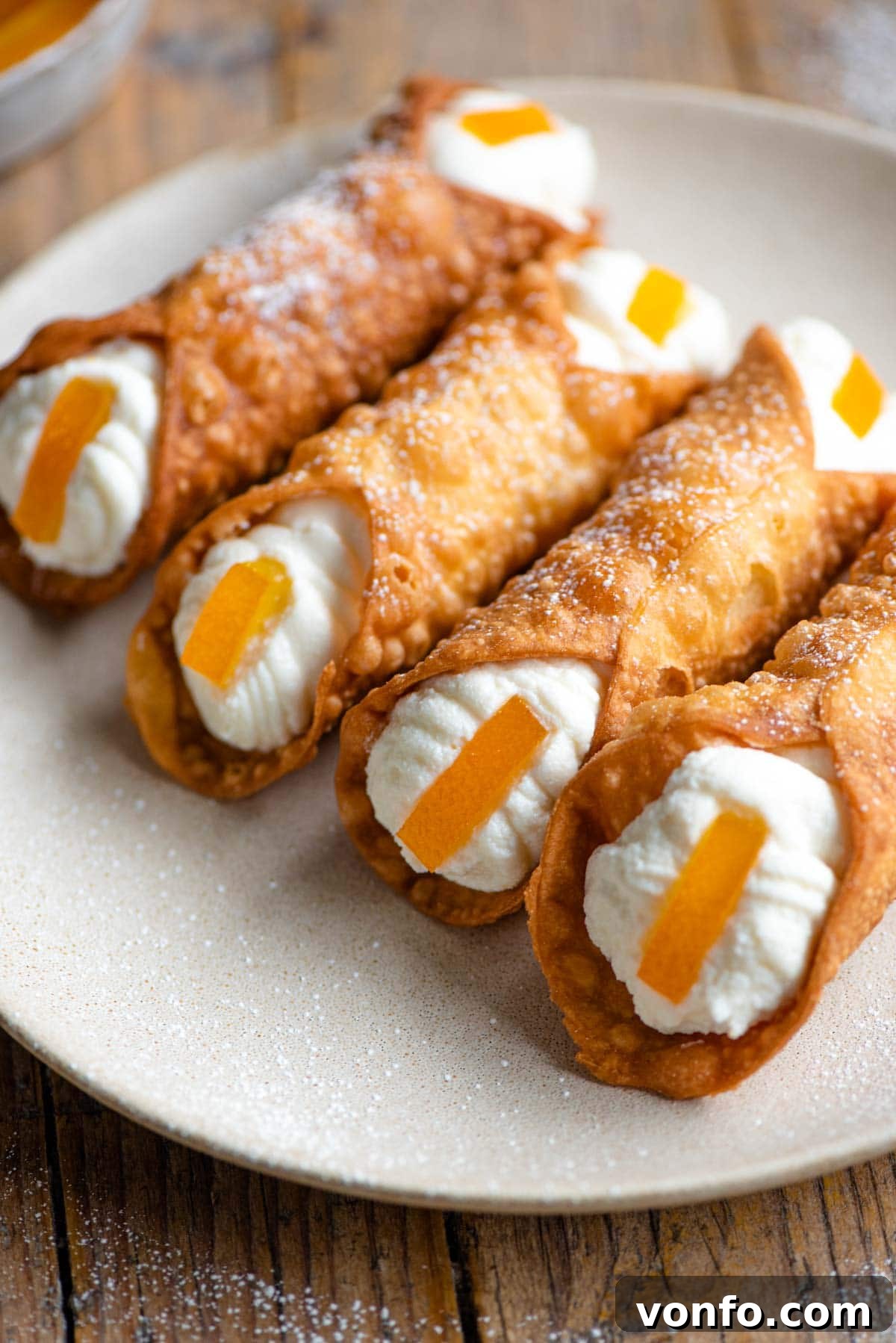 A close-up view of four artfully arranged cannoli, bursting with creamy ricotta filling, showcasing their crispy, golden shells. Some are garnished with candied orange, others with chopped pistachios.