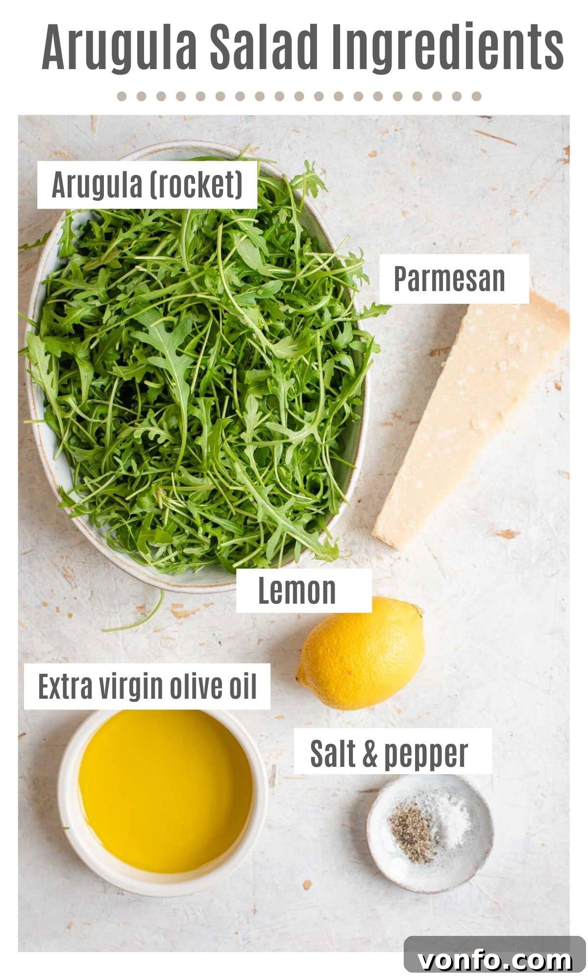 An overhead shot displaying all the fresh ingredients needed to prepare a simple arugula salad: fresh arugula leaves, a wedge of Parmesan cheese, a whole lemon, a bottle of extra virgin olive oil, and a black pepper grinder.