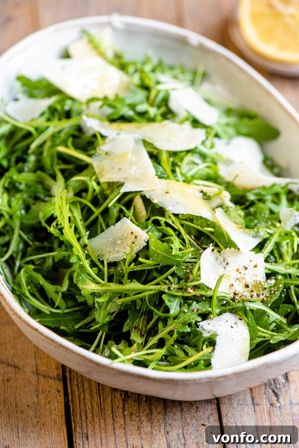 A close-up shot of an arugula salad, highlighting the vibrant green leaves, visible Parmesan shavings, and specks of freshly ground black pepper in a stylish bowl.