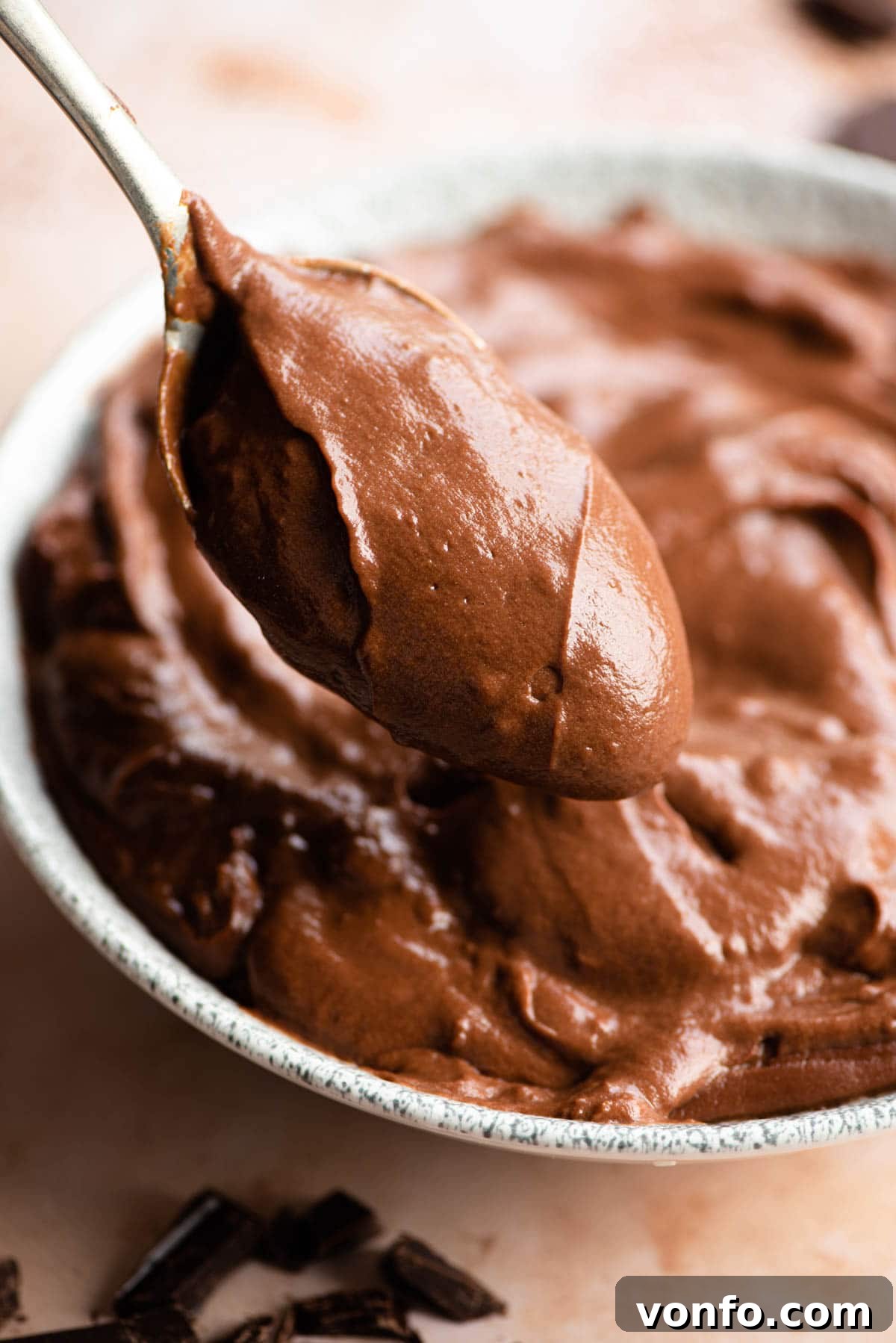 A close up of a spoonful of velvety chocolate pastry cream being lifted from a white bowl, showcasing its smooth texture.