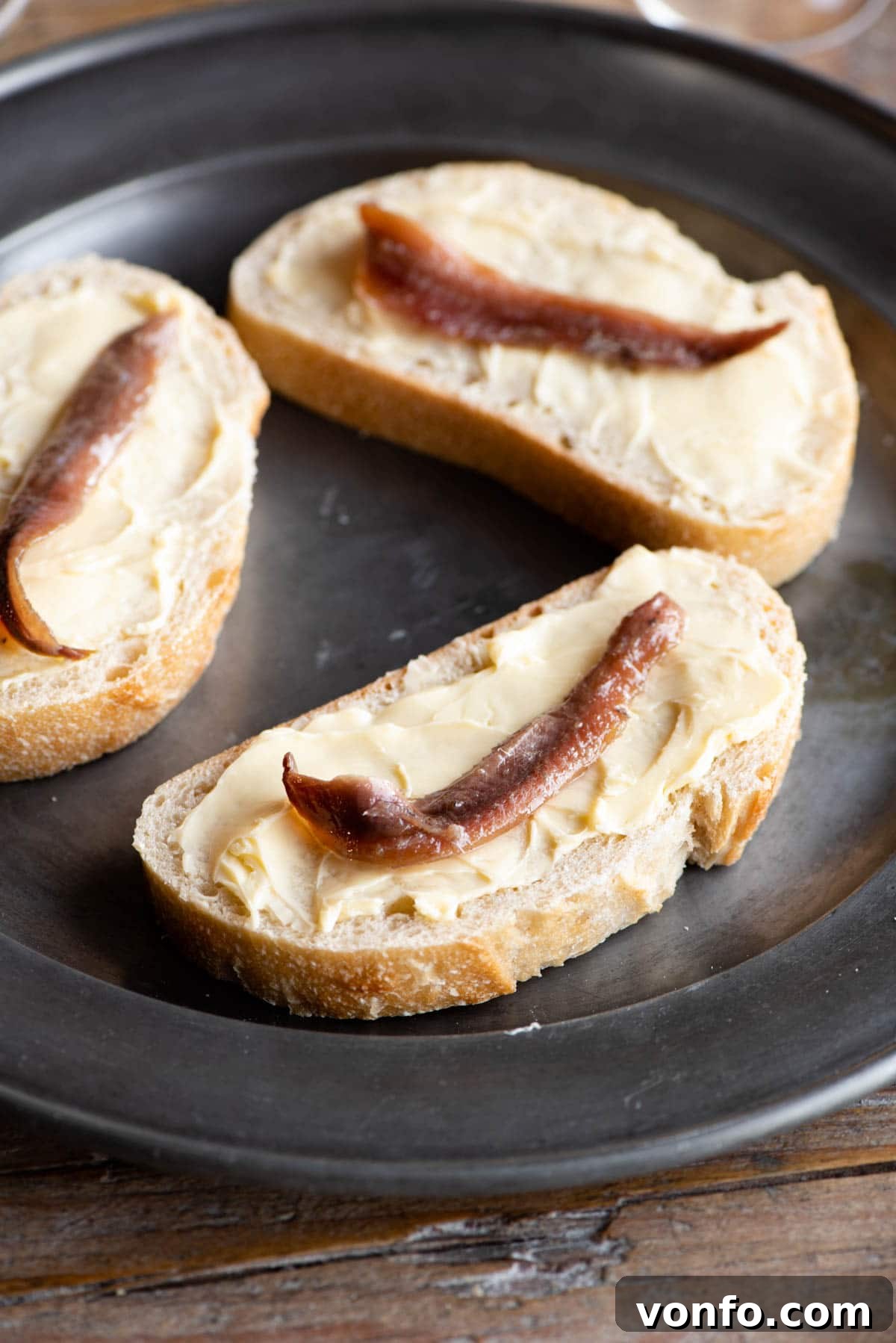 A close-up of a single slice of Pane, Burro e Alice, showing the rich butter and a perfectly placed anchovy fillet on crusty white bread
