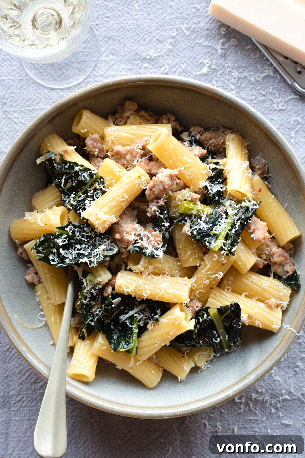 An overhead shot of a bowl of pasta with kale and sausage with a glass of wine at the side, showcasing the rich texture and vibrant colors of the dish.
