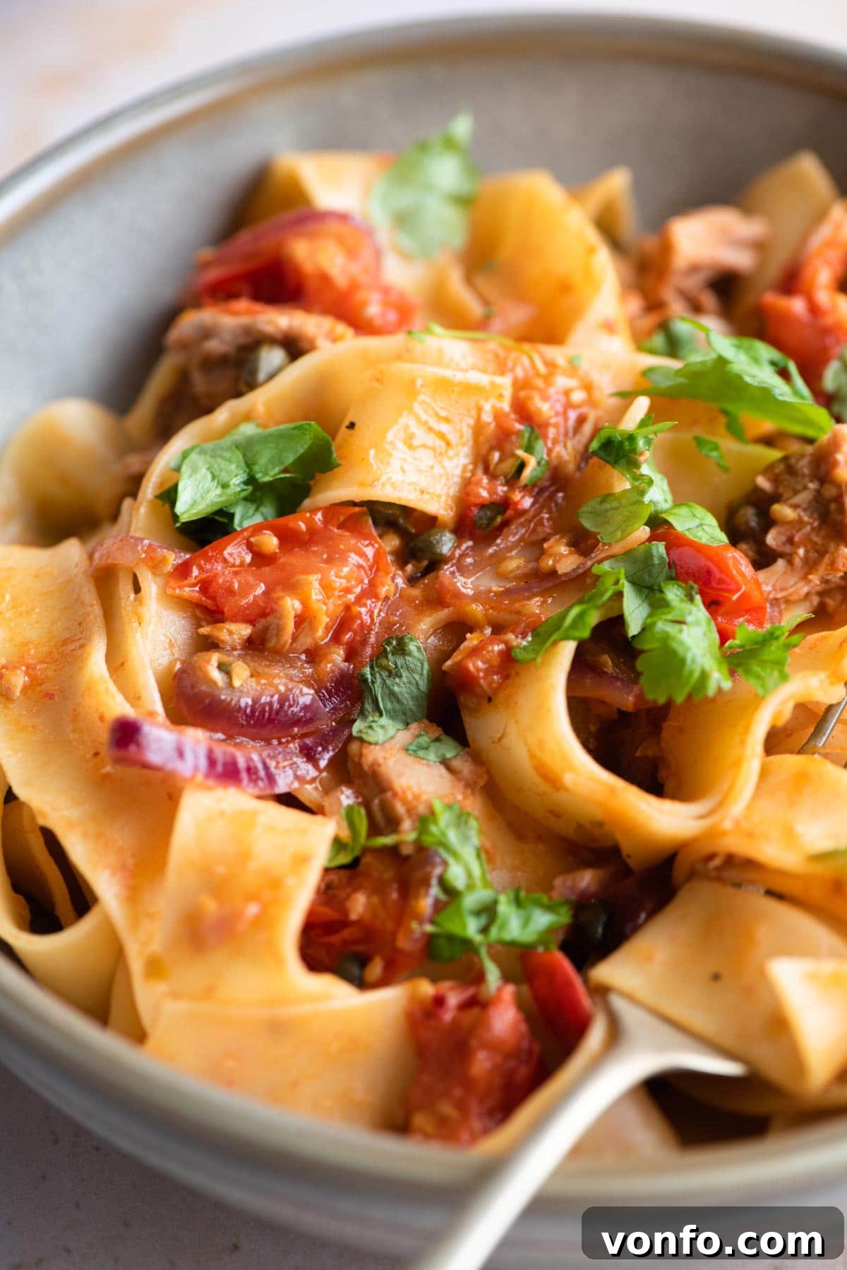 Quick Tuna Pasta with Burst Tomatoes and Briny Capers 6 A close-up photograph of pappardelle pasta generously mixed with tuna, vibrant cherry tomatoes, finely sliced onion, and briny capers.